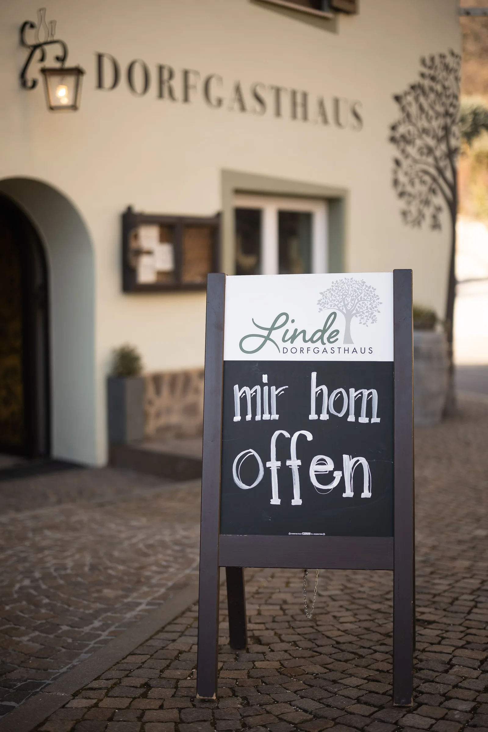 Tafel mit der Aufschrift „mir hom offen“ vor dem Dorfgasthaus zur Linde in Kaltern